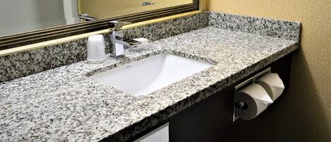 A bathroom with a granite countertop, a white sink, a mirror with a gold frame, and a paper towel dispenser.