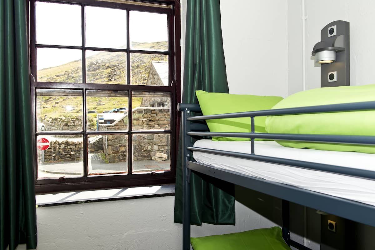 A bunk bed with green bedding, a window with a view of a stone wall and a car, and a wall-mounted light fixture.