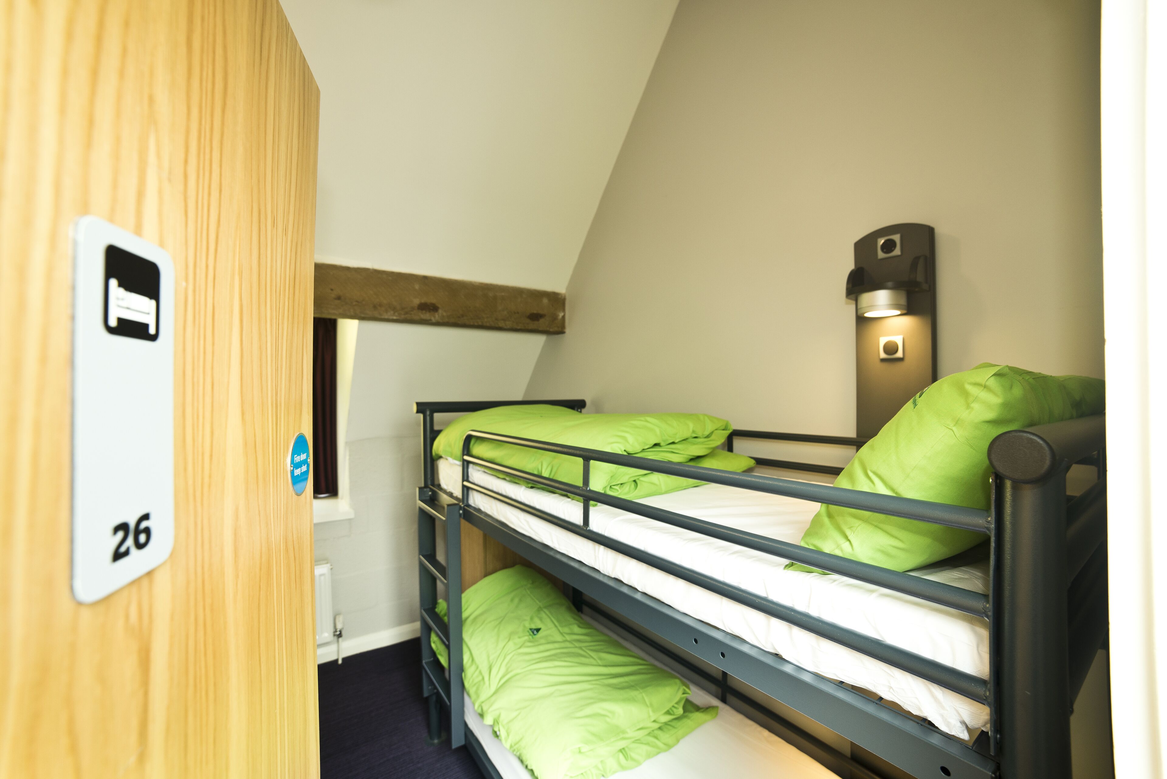 A bunk bed with green bedding, a wooden door with a number, and a wall-mounted light fixture.