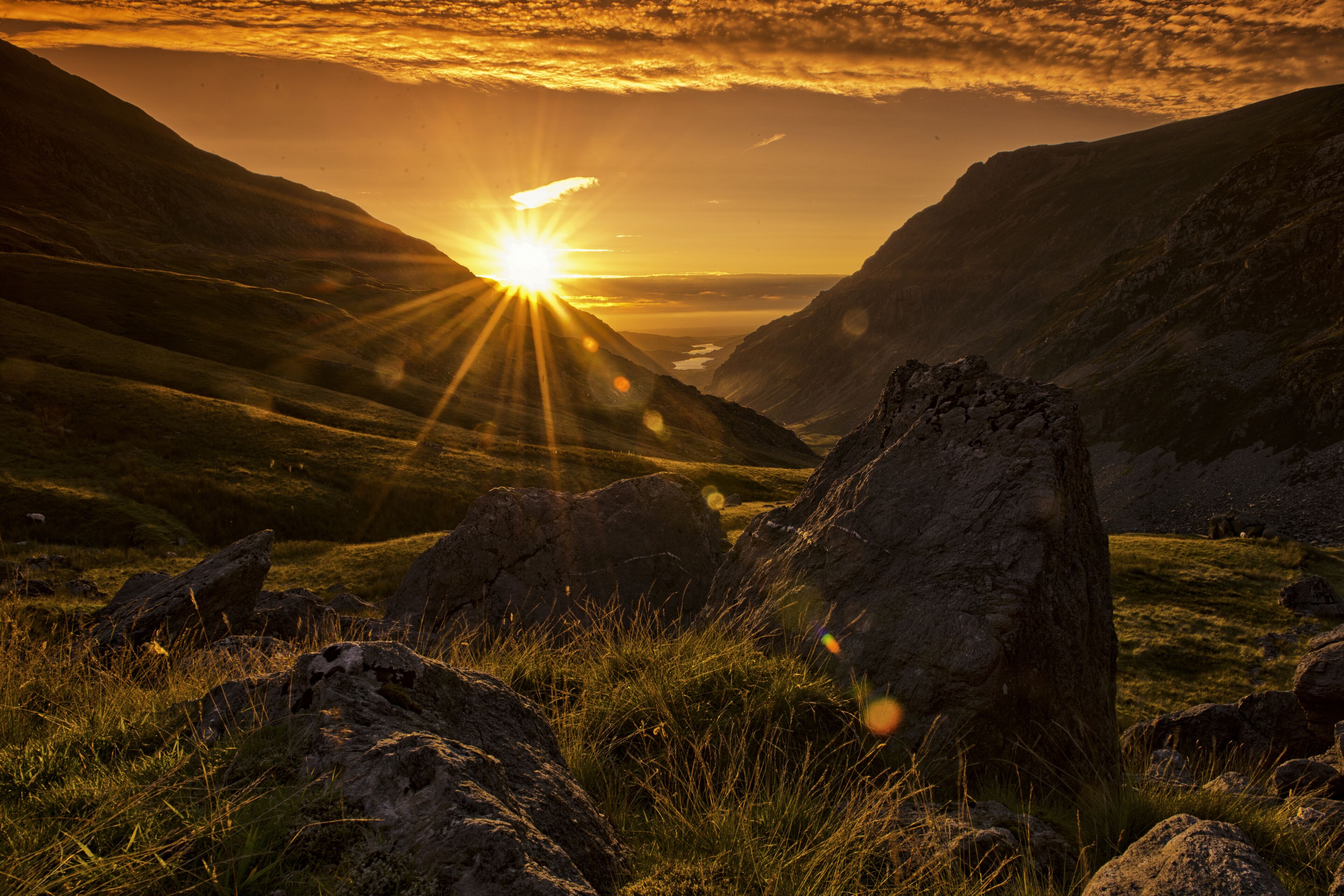 Un paysage montagneux, avec un soleil couchant derrière des formations rocheuses et des collines couvertes d’herbe.