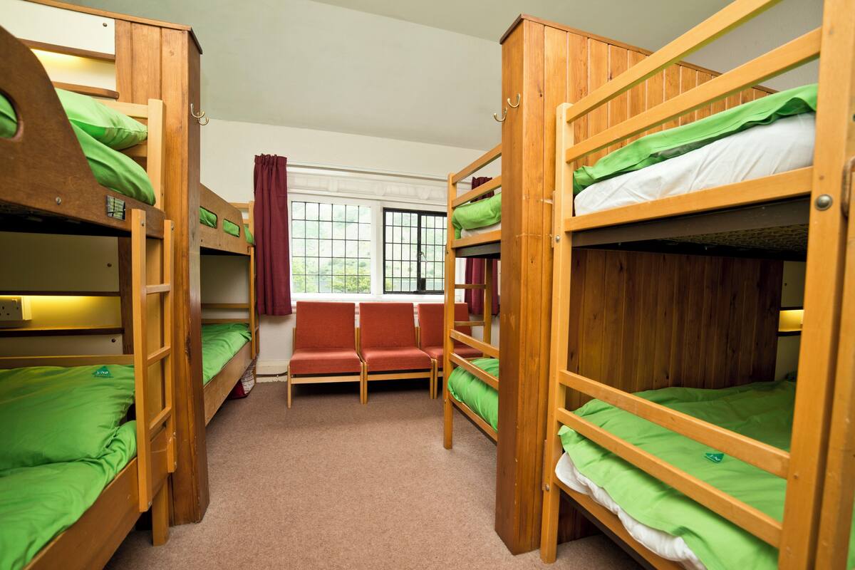A bunk room with wooden bunk beds, a red bench, and a window with curtains.