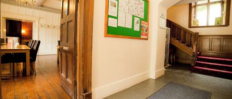 A hallway with a wooden staircase, a bulletin board with notices, and a dining area with a table and chairs.