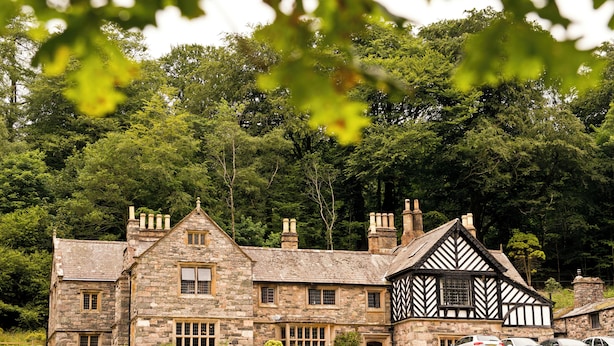 Un bâtiment en pierre, dont l’architecture combine des éléments traditionnels et des éléments de style Tudor, avec de nombreuses cheminées et une grande pelouse à l’avant.