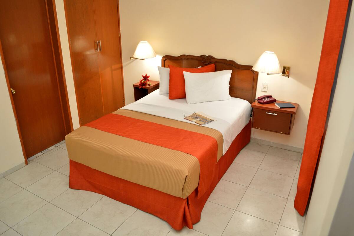 A hotel room with a bed, nightstands, a wooden wardrobe, and a red curtain.
