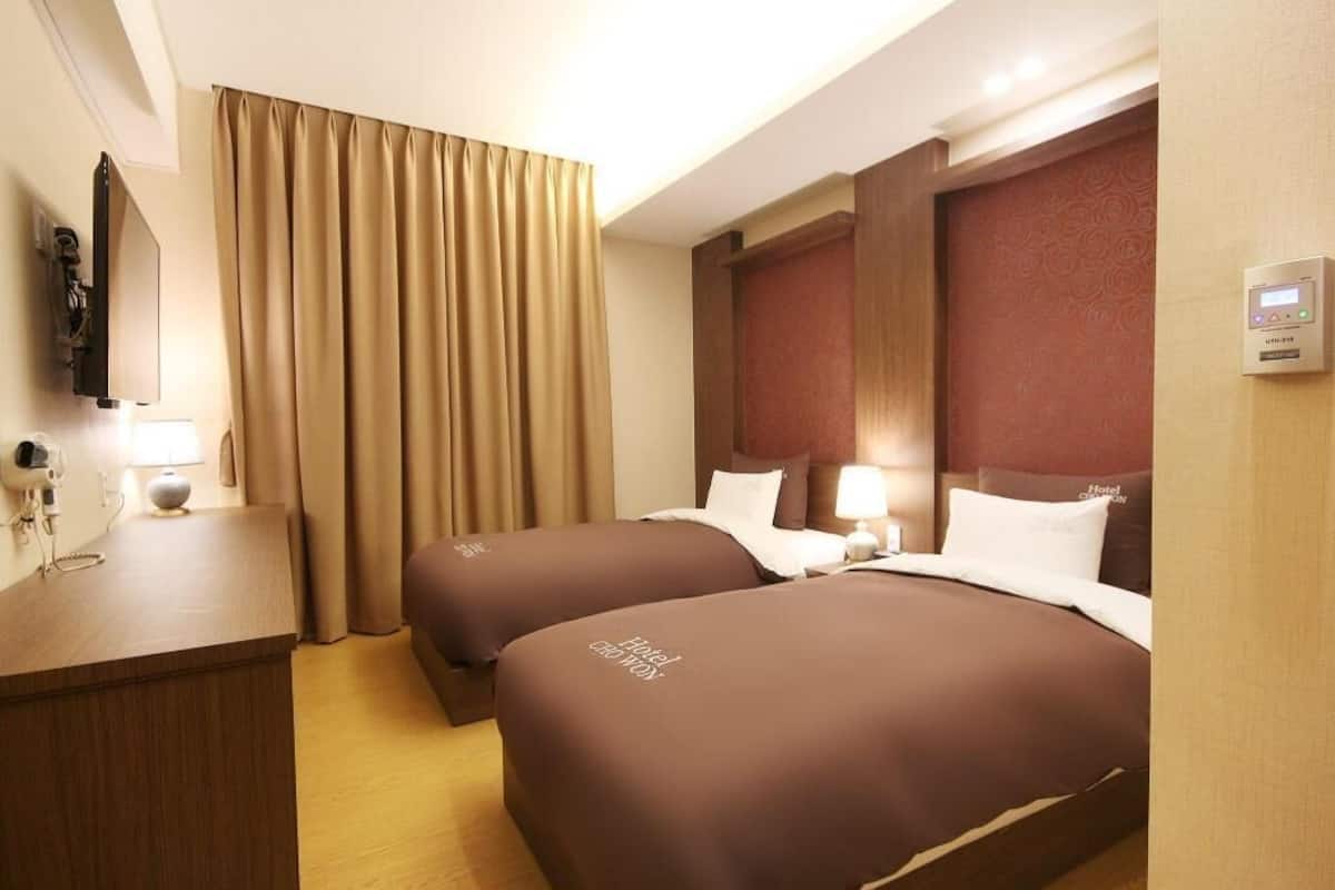 A hotel room with two beds, a desk, a mirror, a TV, and a control panel on the wall.