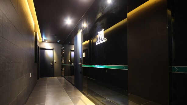 Um corredor moderno com piso escuro, iluminado por luzes no teto, que leva a uma área de recepção com porta de vidro e uma placa em cima.