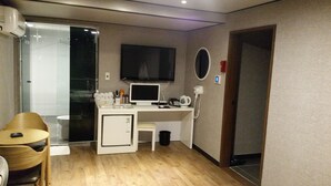 Quarto de hotel compacto com uma escrivaninha de madeira, um computador, uma TV, um frigorífico pequeno, uma cadeira e uma mesa de cabeceira.