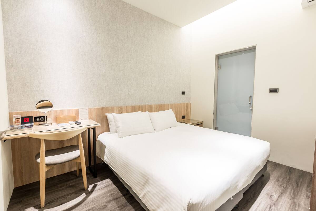 A hotel room with a bed, a desk, a chair, a nightstand, and a door.