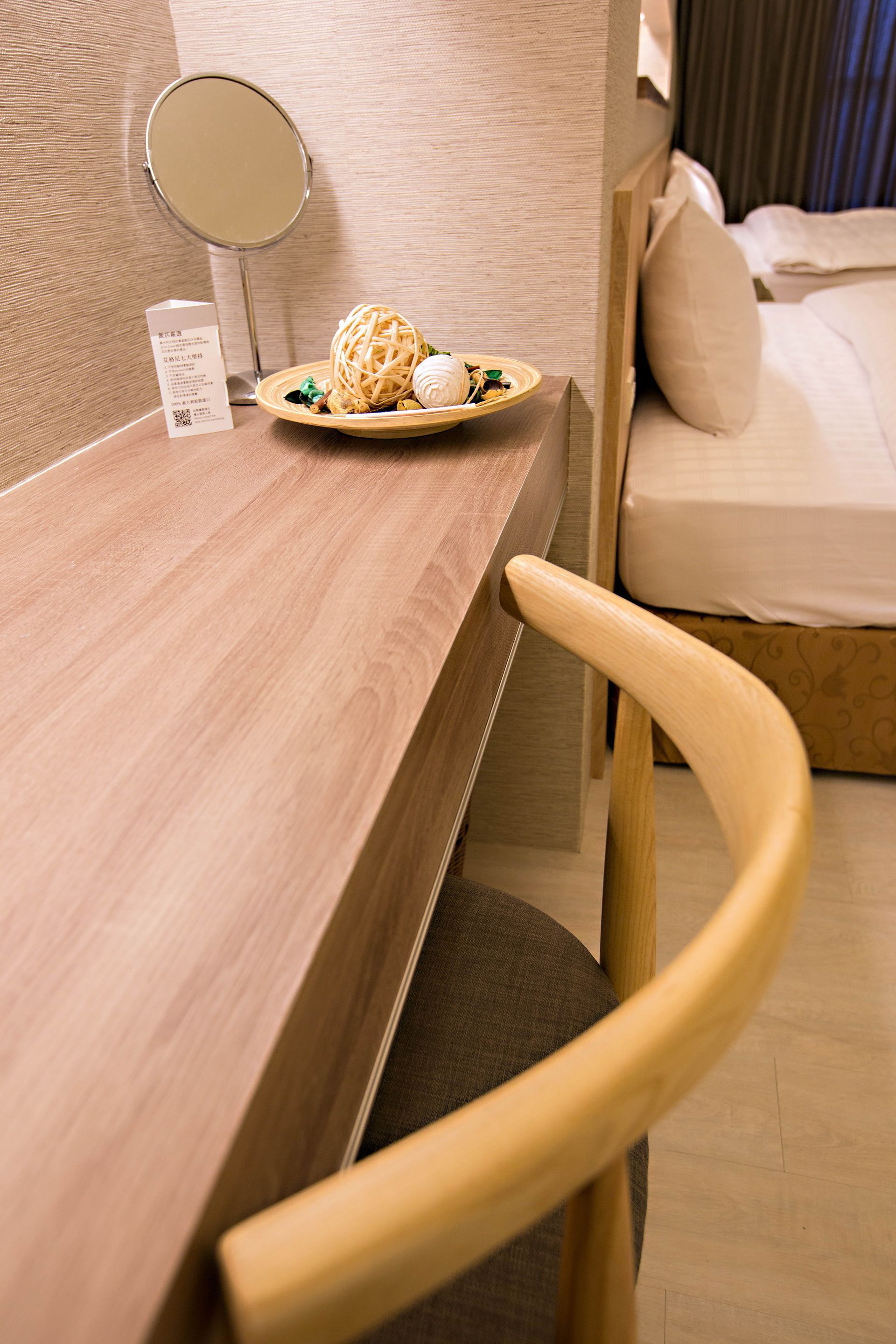 Une table en bois avec une assiette de nourriture et un miroir. On aperçoit, à l’arrière-plan, un lit avec un matelas et des oreillers blancs.