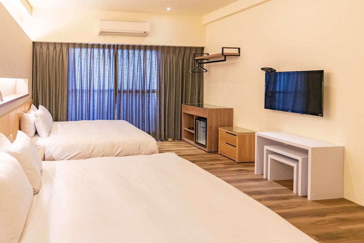 A hotel room with two beds, a TV mounted on the wall, a desk, and a small seating area.