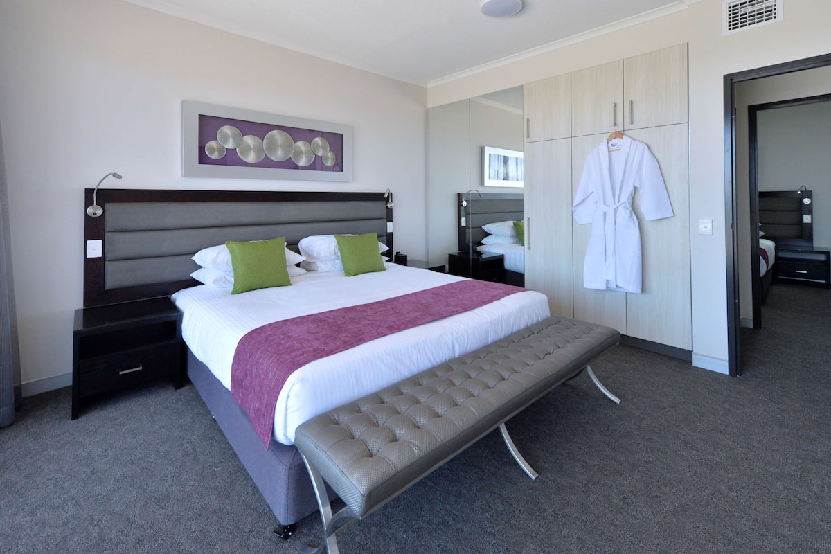 A modern hotel room with a large bed, a bench, a wardrobe, and a mirror.