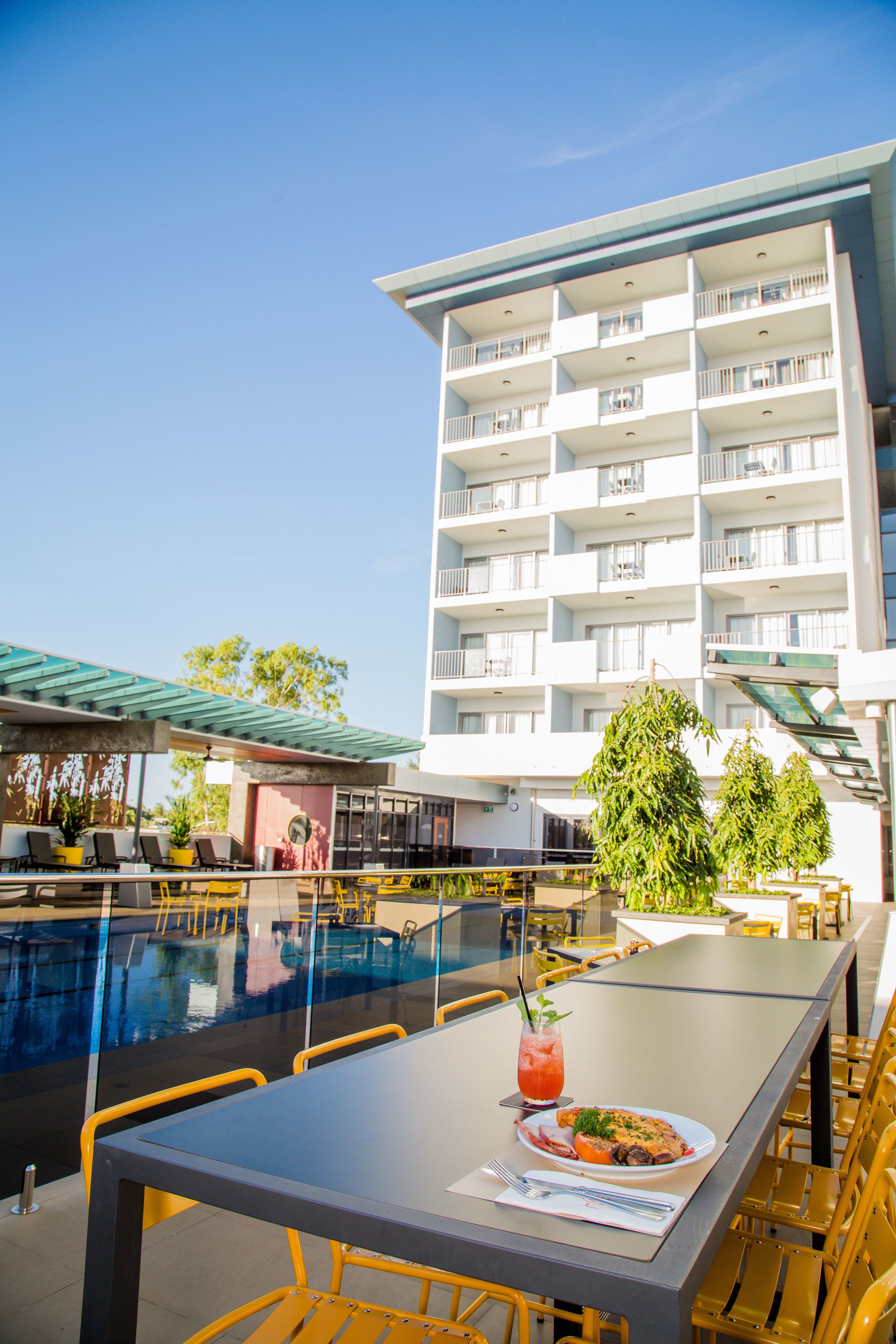 Ein Essbereich am Pool mit Tisch, Stühlen und einem Getränk. Im Hintergrund ein Gebäude mit Balkonen.