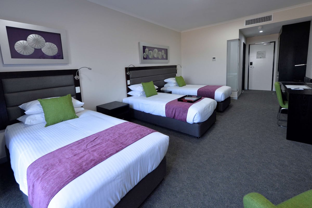 A hotel room with three beds, each with a different colored pillow, a desk with a chair, and artwork on the walls.
