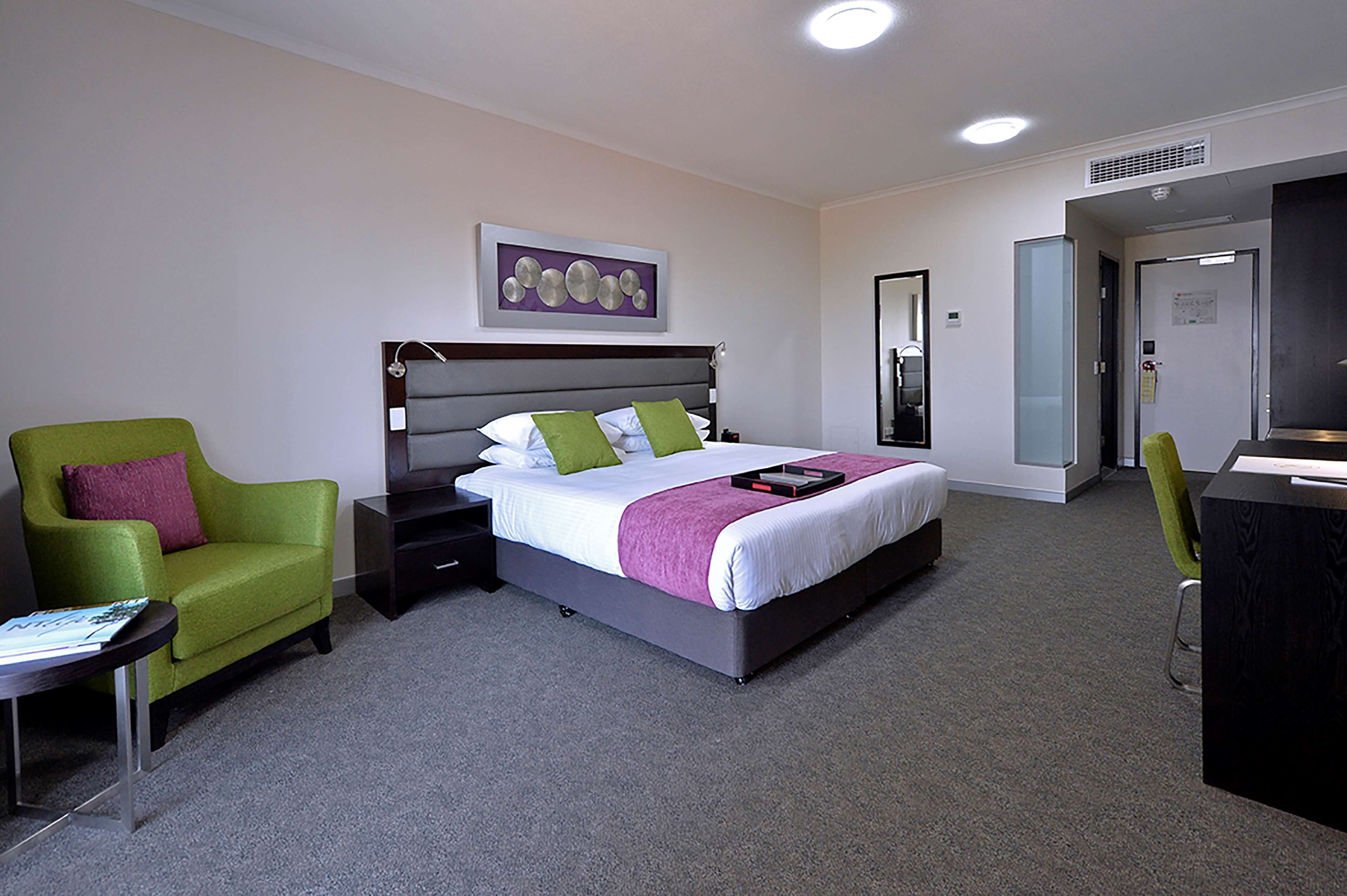 A hotel room with a bed, a green armchair, a desk, and a mirror.