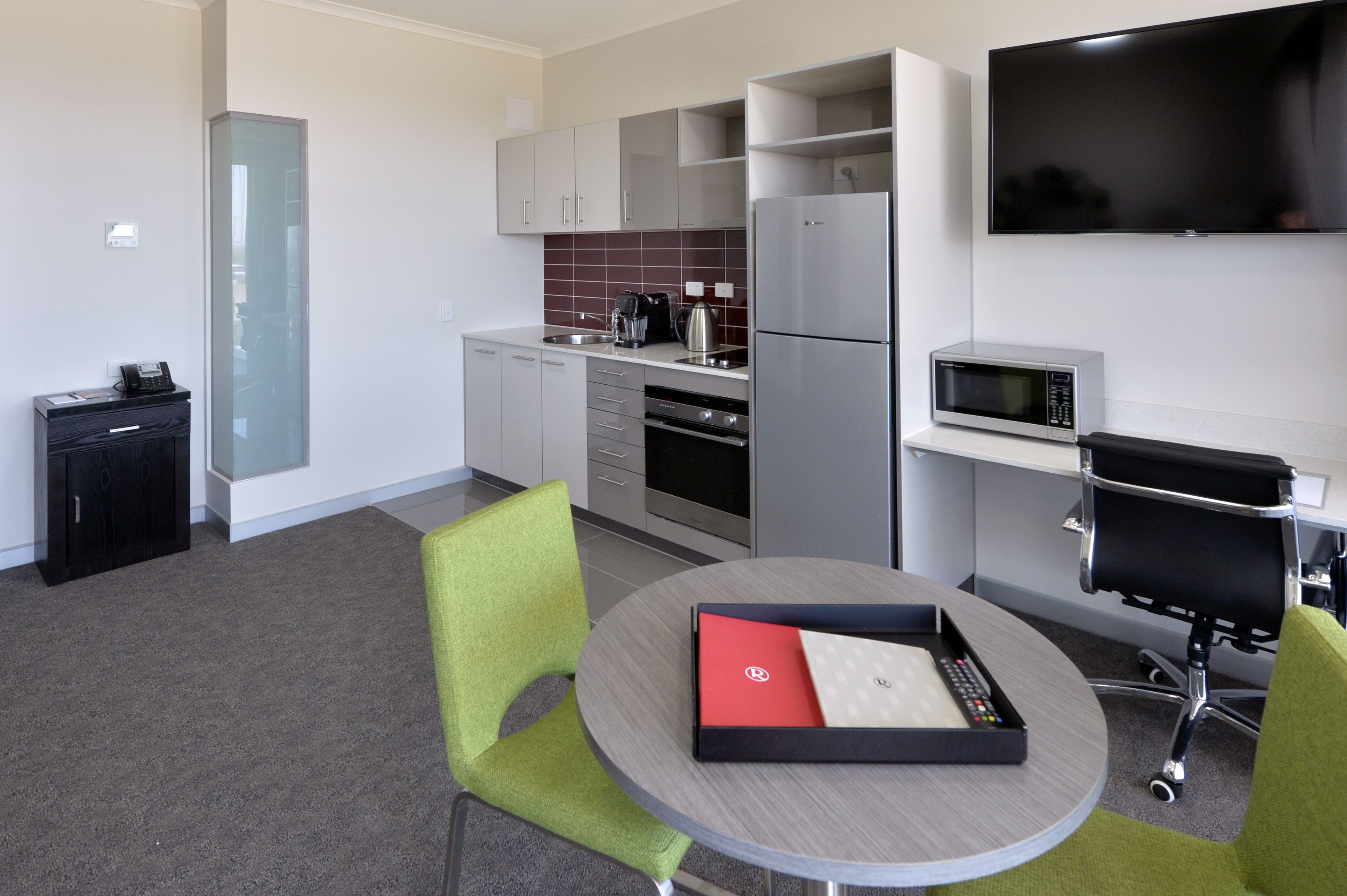 A modern kitchen with a refrigerator, microwave, and coffee maker. There is a dining table with chairs and a TV mounted on the wall.