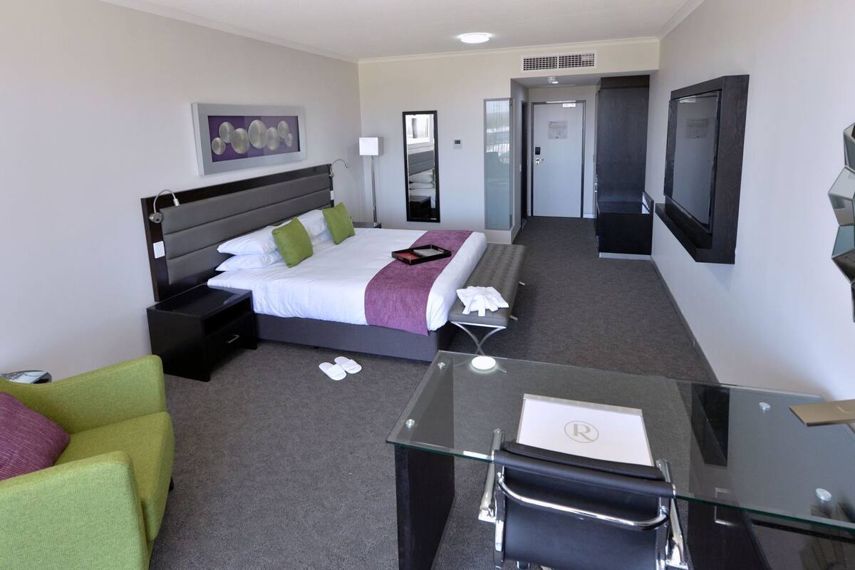 A hotel room with a bed, bedside tables, a desk with a chair, a sofa, a TV, and a door.