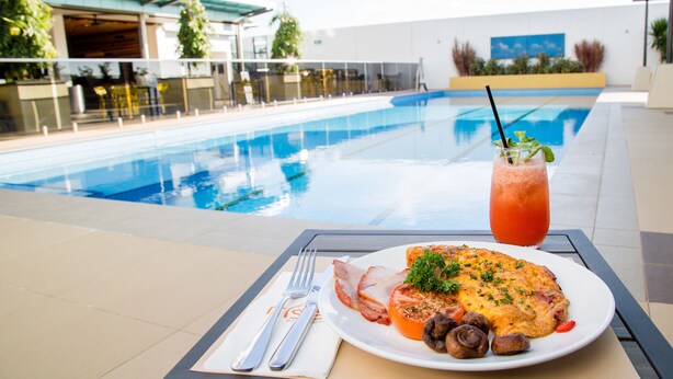 A dining table with a plate of food, a drink, and a view of a swimming pool.