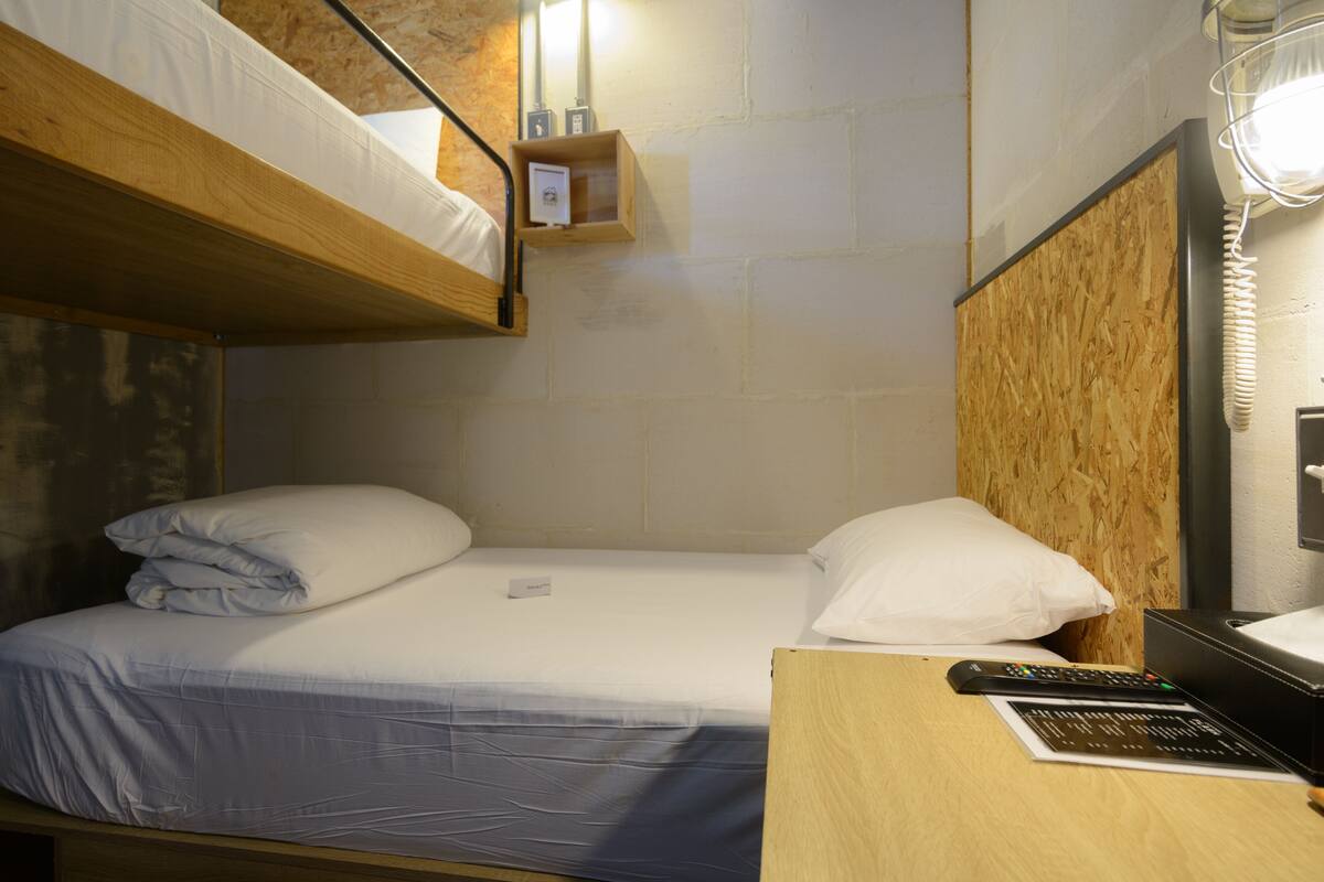 Uma cama de solteiro com cabeceira de madeira, uma mesinha de cabeceira com um controle remoto e uma pequena prateleira com um cartão.
