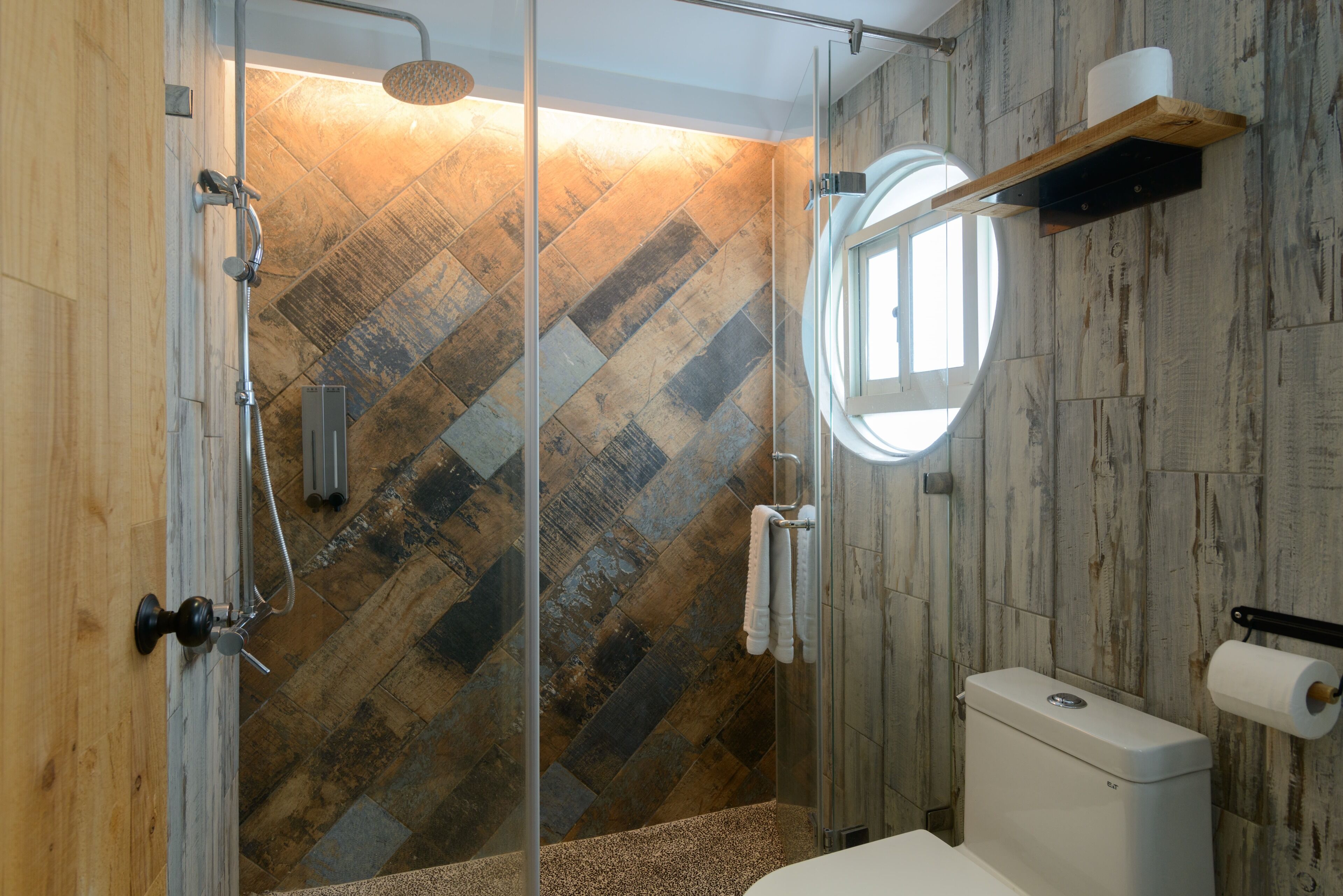Ein modernes Badezimmer mit Wandgestaltung aus Fischgrätmusterfliesen, einer Duschkabine aus Glas, einer Toilette und einem Holzregal mit Handtuchhalter.