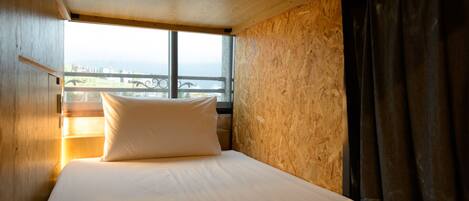 Una cama individual con colchón y almohada blancos, una ventana con vista a edificios y una cortina a la derecha.