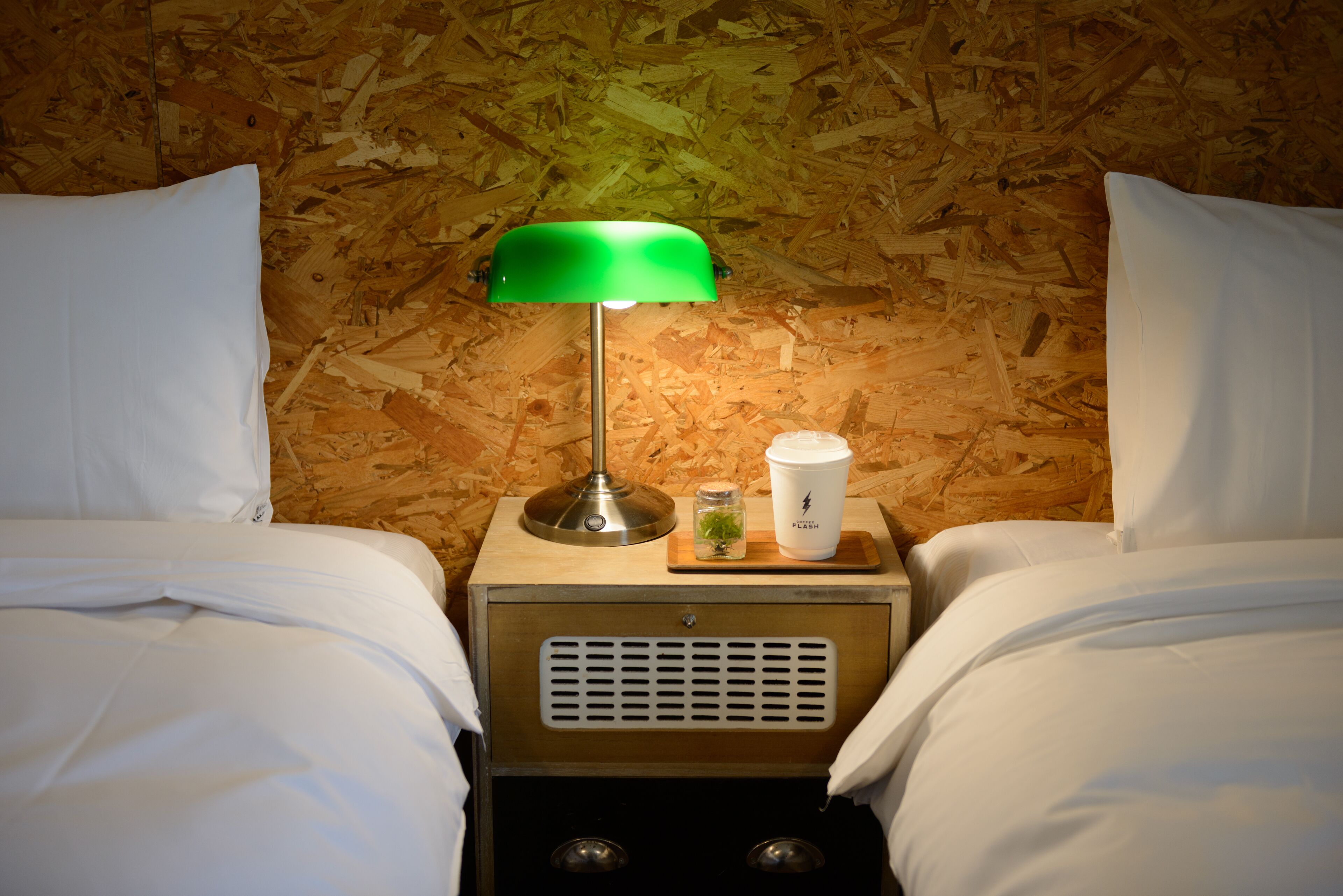 A hotel room with two beds, a nightstand with a lamp, a cup, and a small plant.