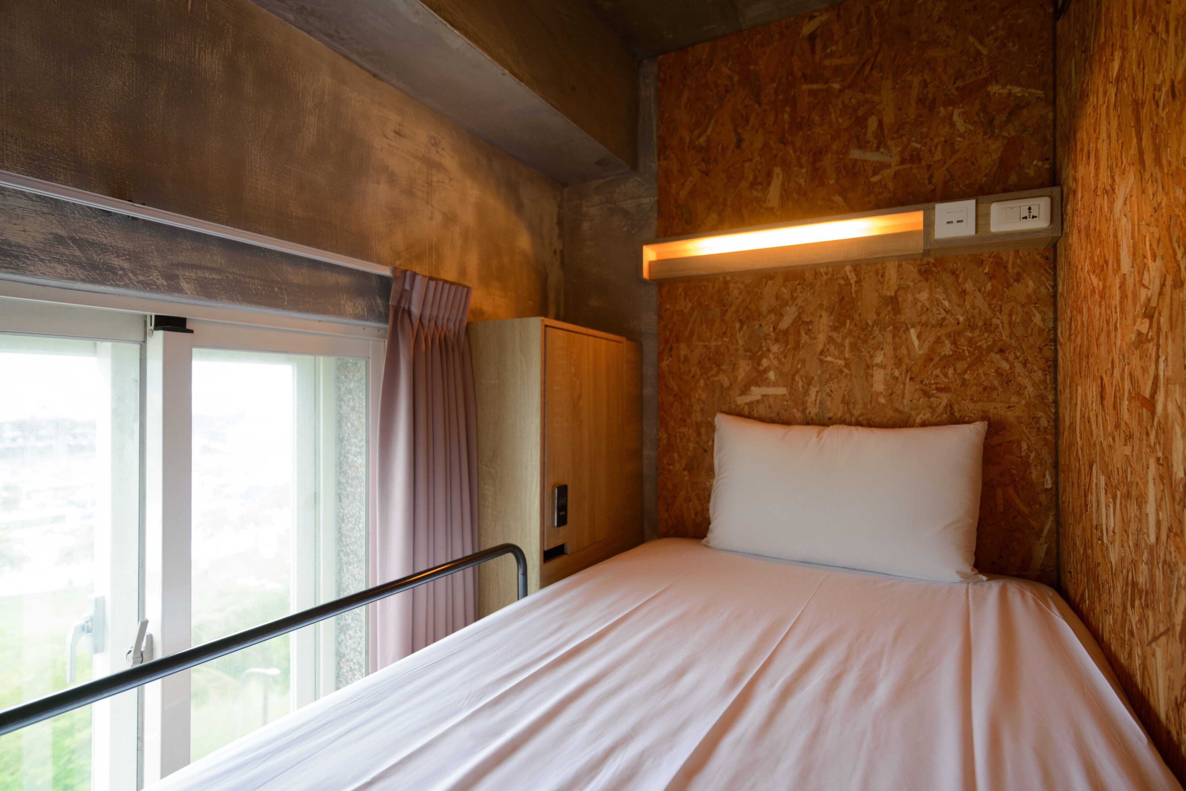 Habitació petita amb un llit individual, paret de fusta, finestra amb cortines i una tauleta de nit.