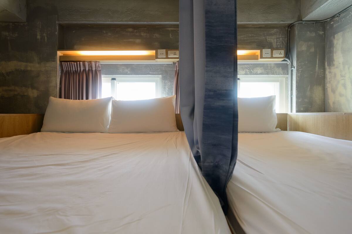 Uma cama de casal com lençóis brancos e duas almofadas, uma janela com cortinas e um ponto de luz fixado na parede.
