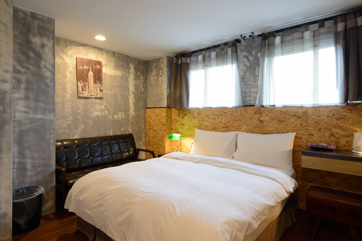 Quarto de hotel com cama, cabeceira, mesa de cabeceira, luminária e janela com cortinas.