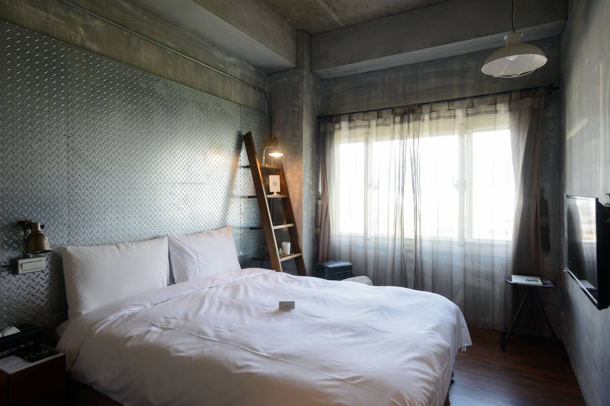 Quarto com cama, mesa de cabeceira, cômoda, escada, janela com cortinas leves e luminária suspensa.