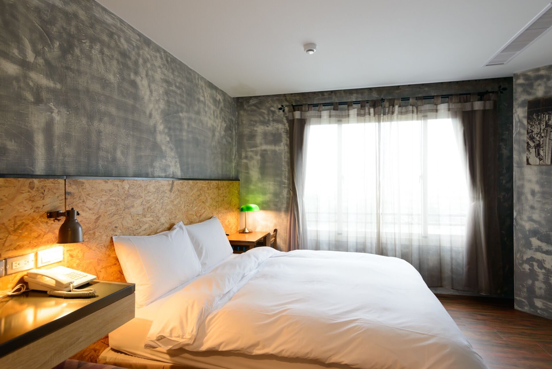 A modern hotel room with a large bed, a bedside table with a lamp, a telephone, and a headboard with a textured wall.
