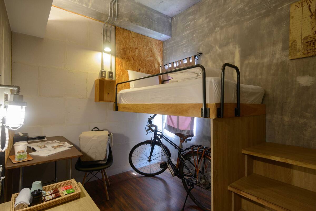 Quarto compacto com uma cama beliche, uma bicicleta, uma mesa pequena com luminária e uma prateleira na parede.