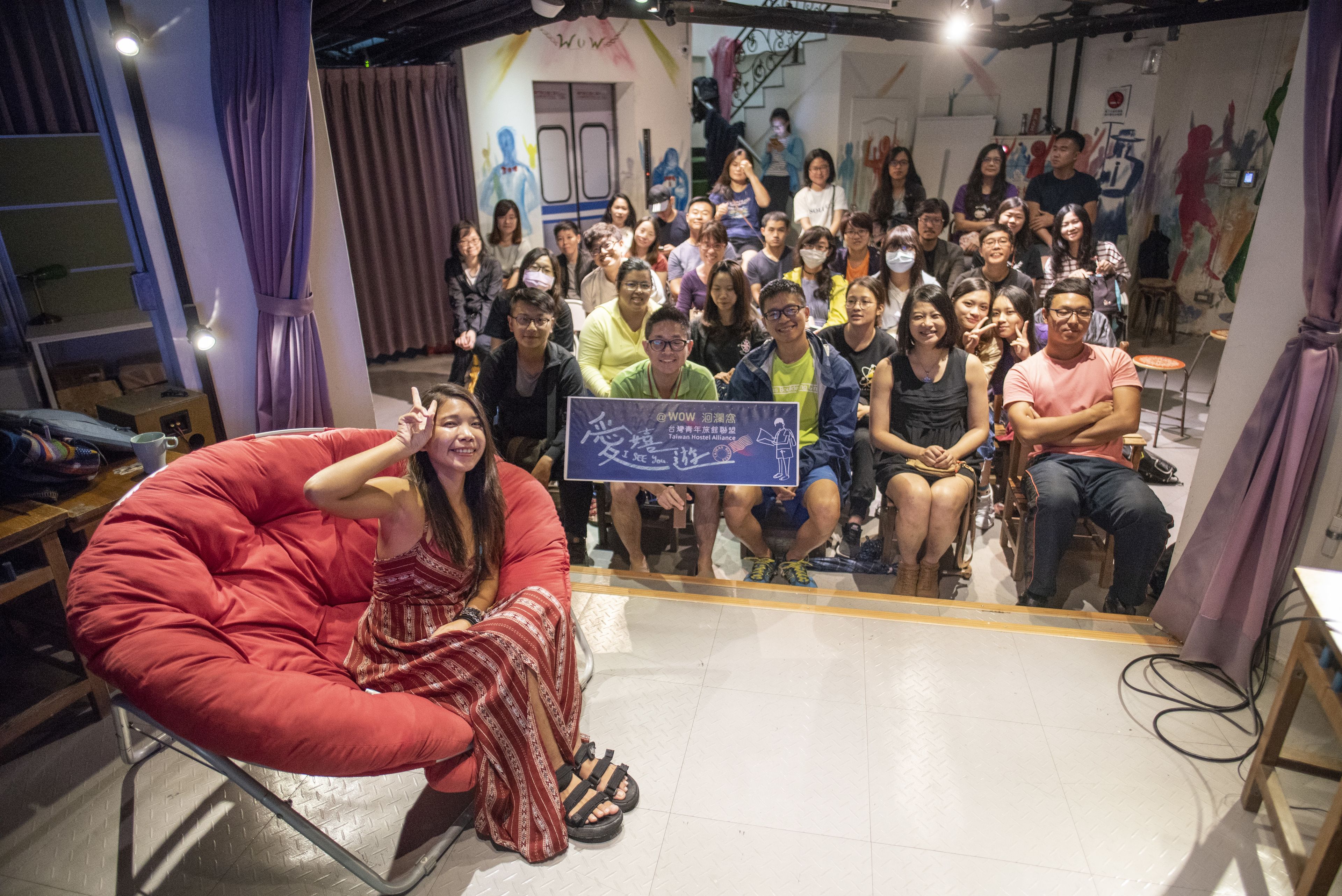 A group of people seated in a room with a stage, a banner, and a curtain.