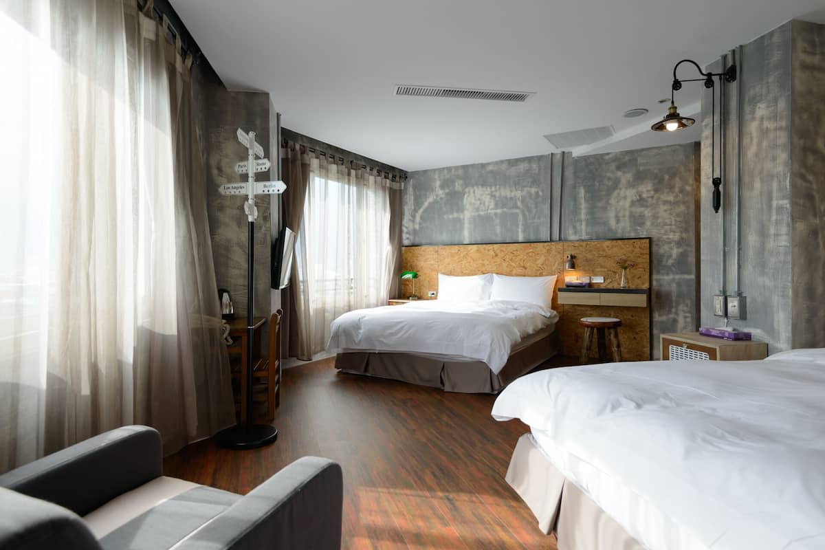 Quarto de hotel com duas camas, uma escrivaninha, uma cadeira, um abajur e uma janela grande com cortinas.