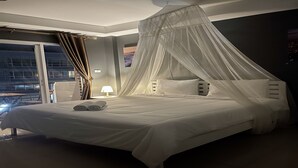 A modern hotel room with a large bed, a canopy, and a night view of the city.