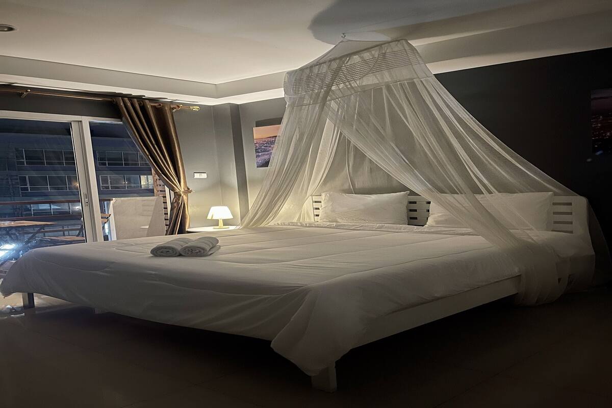 A modern hotel room with a large bed, a canopy, and a night view of the city.