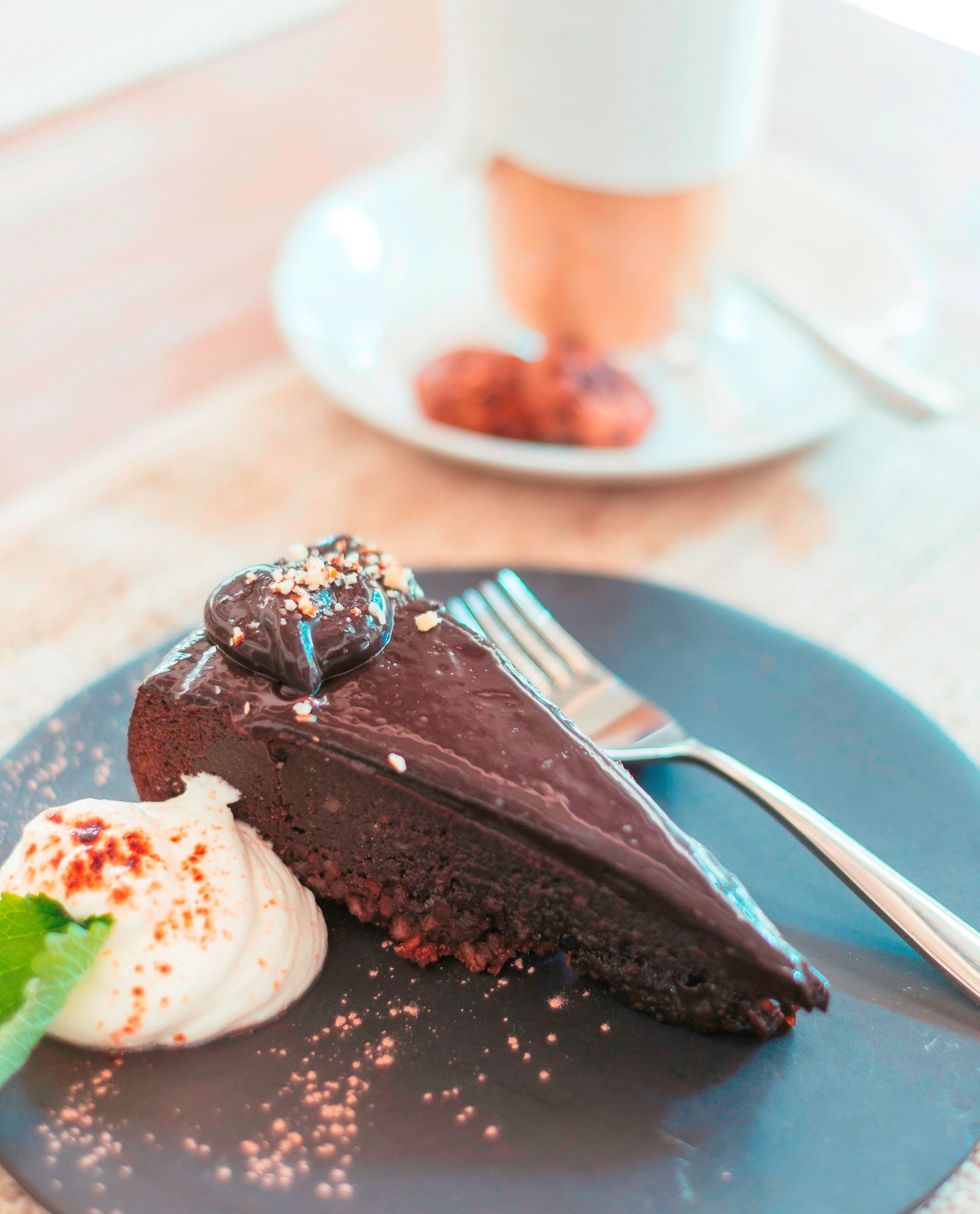 Un plato de postre con pastel de chocolate y crema batida, un tenedor y una taza al fondo.