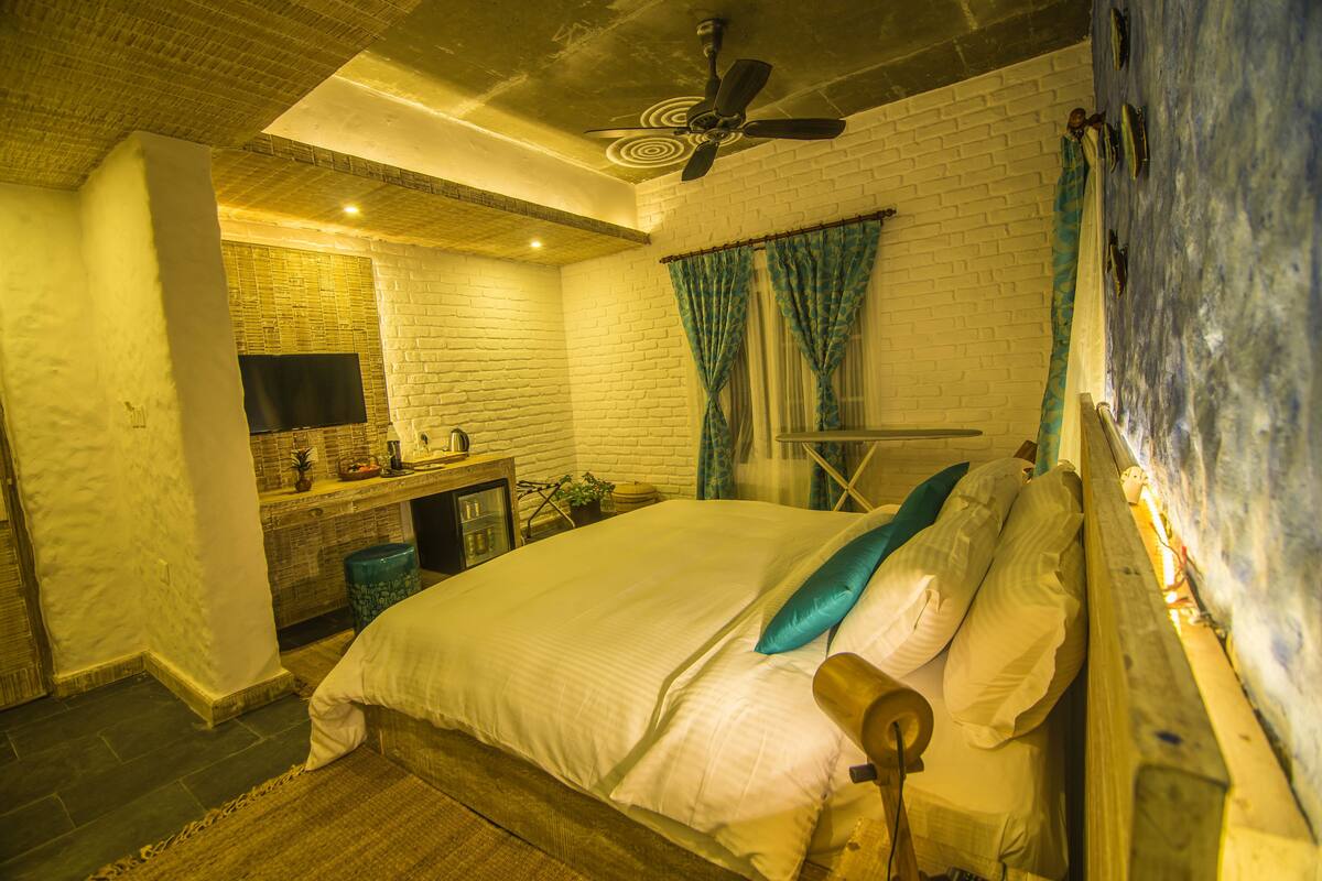A bedroom with a bed, a desk with a flat-screen TV, a ceiling fan, and a brick wall.