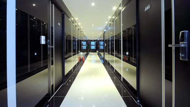 Un couloir d’hôtel moderne avec des portes en verre, un sol réfléchissant et un éclairage au plafond.