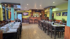 A restaurant with multiple dining tables and chairs, a bar area, and a television screen displaying a menu. - Balthali Village Resort (Balthali)