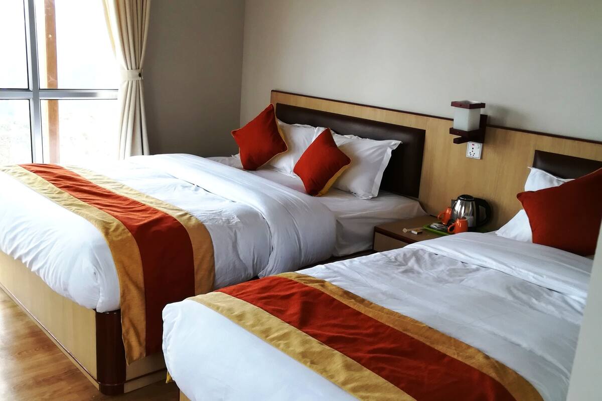 Quarto de hotel com duas camas, cada uma com roupa de cama branca e detalhes vermelhos, cabeceiras de madeira e uma janela com cortinas.