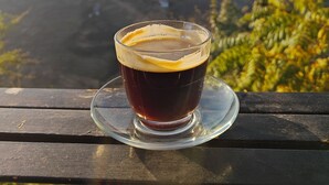 A glass of dark liquid on a clear saucer, placed on a wooden surface with a mountainous landscape in the background. - Balthali Village Resort (Balthali)