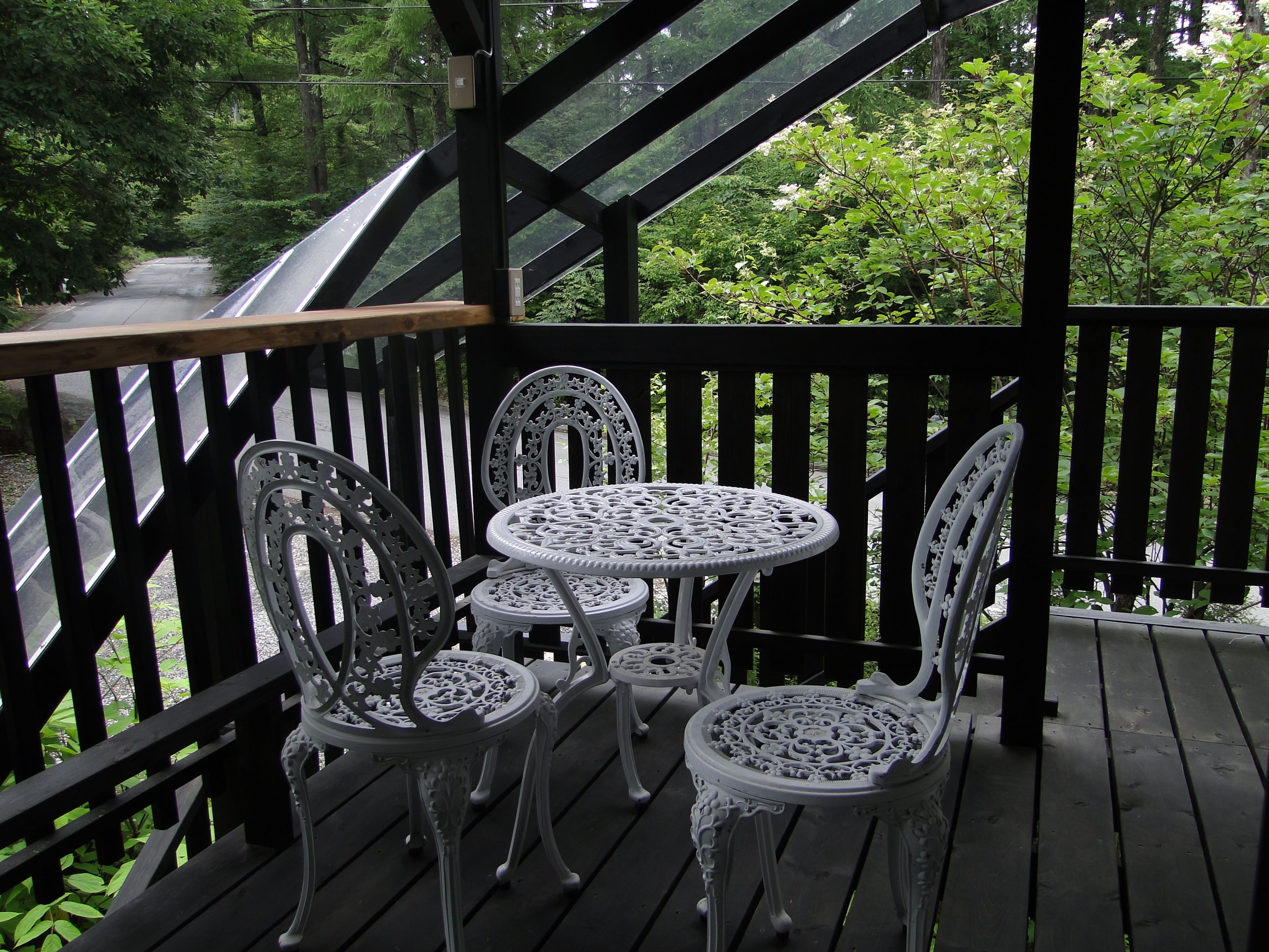 Una terraza de madera con sillas de metal decorativas y una mesa, con vistas a una carretera y abundante vegetación.