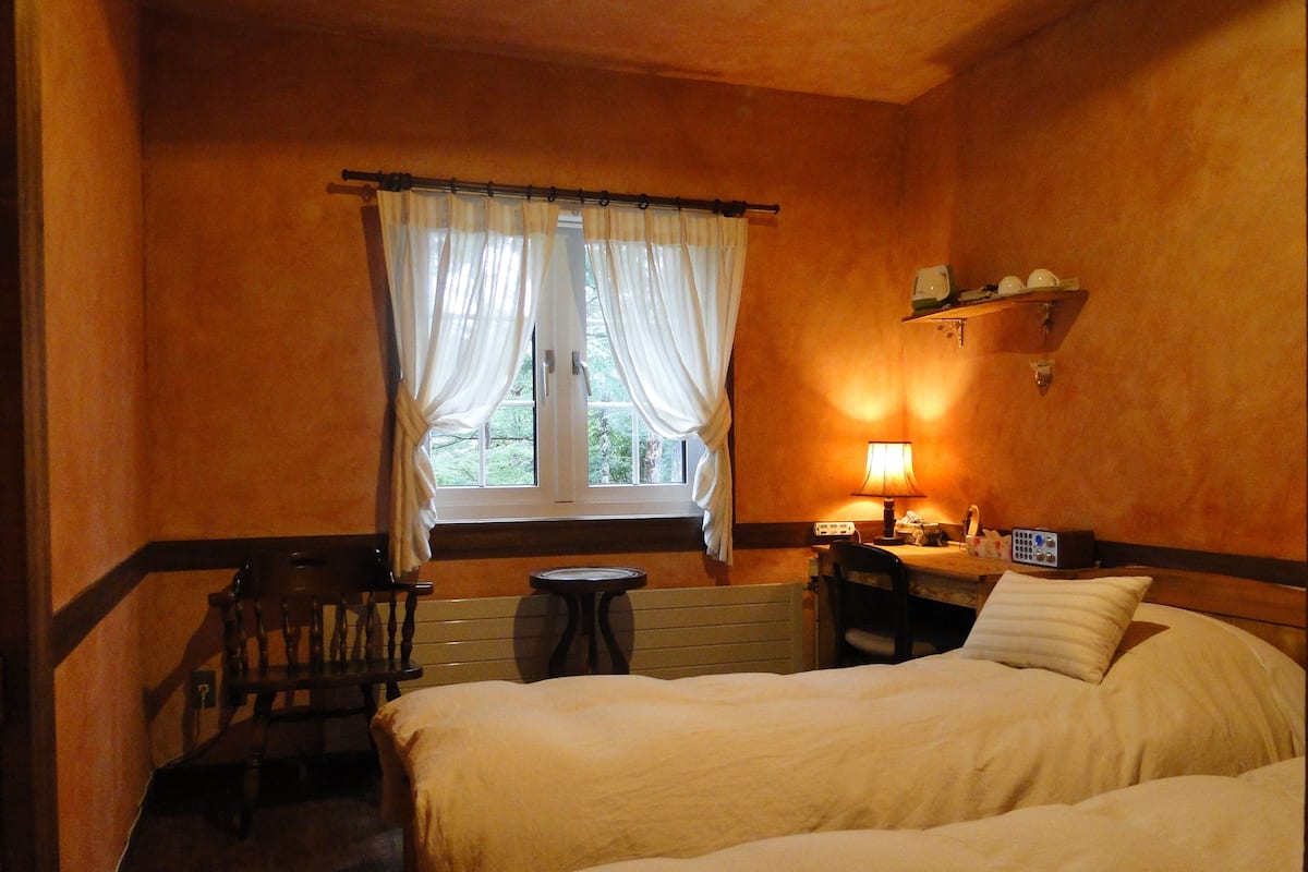 Ein Hotelzimmer mit zwei Betten, einem kleinen Tisch, einem Stuhl, einem Fenster mit Vorhängen, einer Nachttischlampe und einem Regal mit Gegenständen.