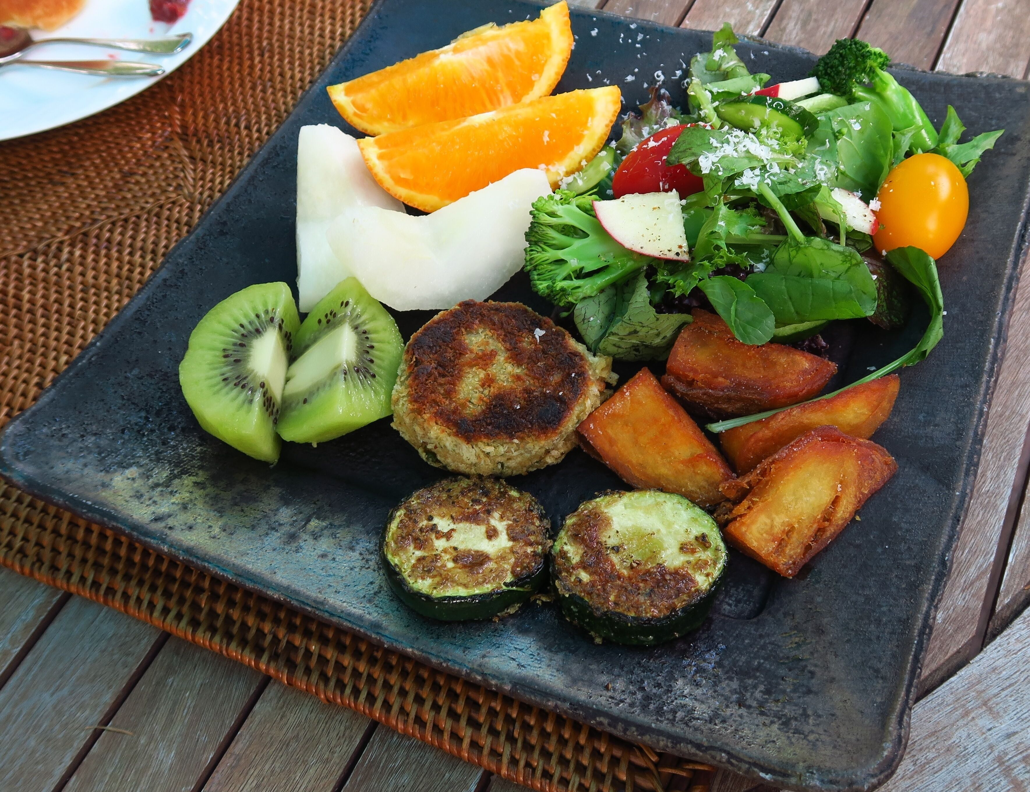 Une assiette carrée contenant divers aliments, notamment des tranches de kiwi, des courgettes grillées, des pommes de terre rôties, une salade composée de jeunes pousses et de tomates cerises, ainsi qu’une tranche de pain blanc.
