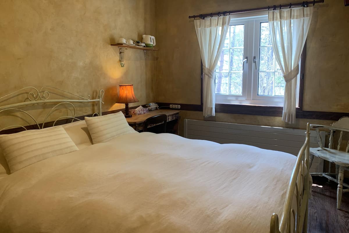 Ein Schlafzimmer mit einem großen Bett, einem Nachttisch mit Lampe, einem Fenster mit Vorhängen und einem Regal mit Tassen und einer kleinen Pflanze.