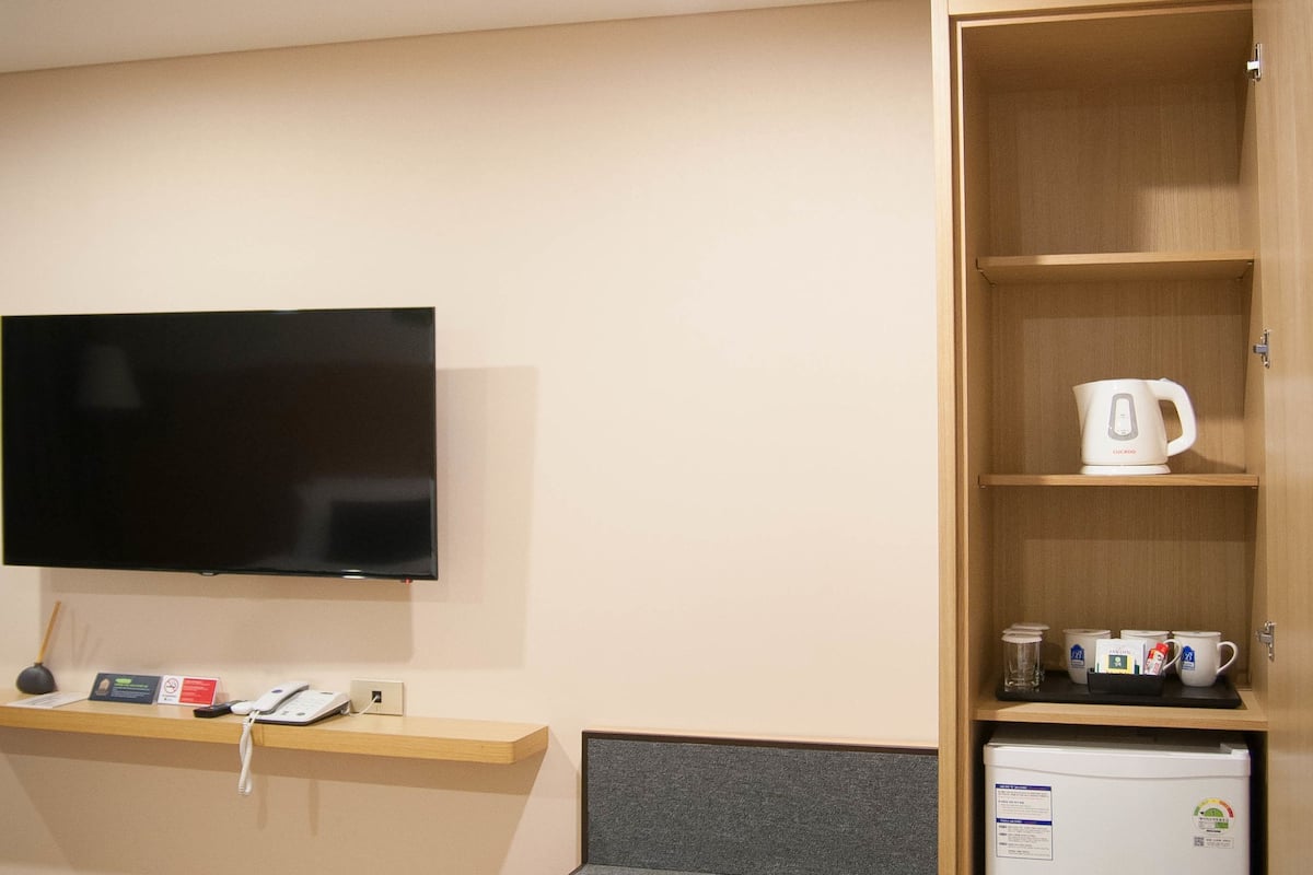 A hotel room with a flat-screen TV mounted on the wall, a small refrigerator, a coffee maker, and a shelf with a phone and a small table.