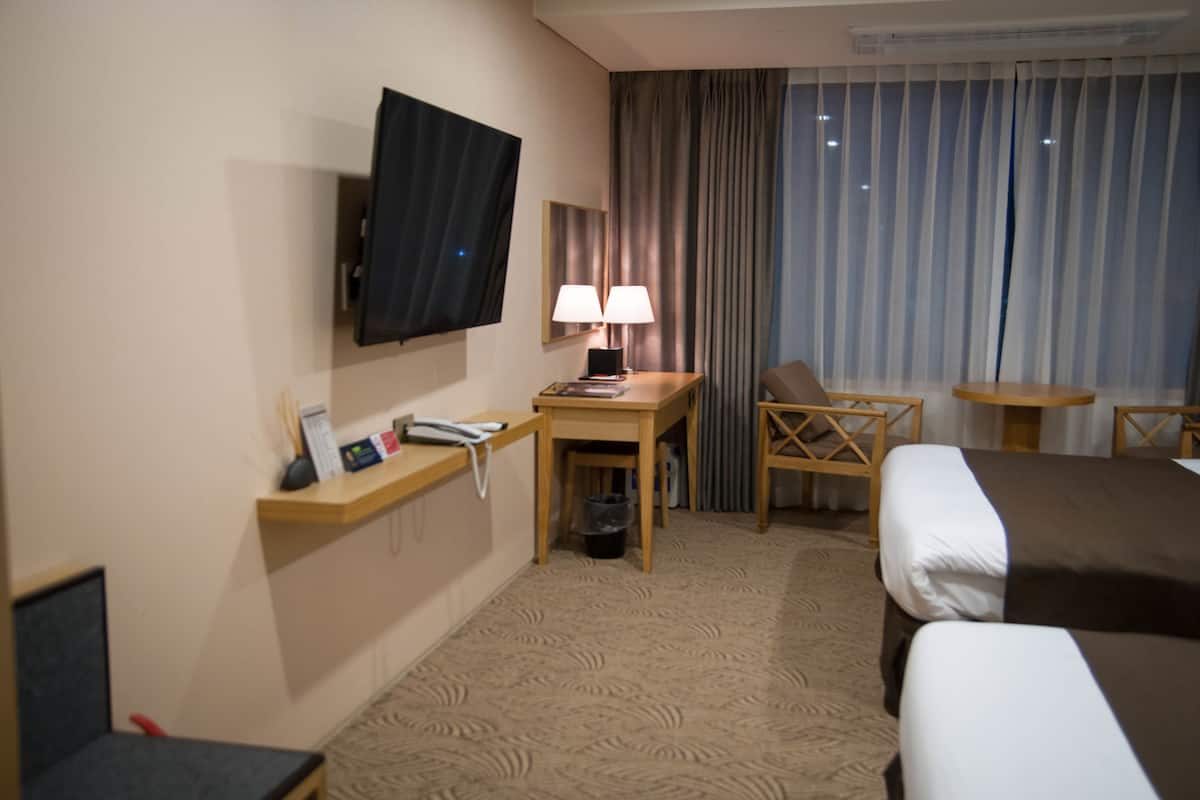 A hotel room with a bed, a desk with a lamp, a TV mounted on the wall, and a window with curtains.