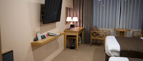 Habitación de hotel con cama, escritorio con lámpara, televisión montada en la pared y ventana con cortinas.