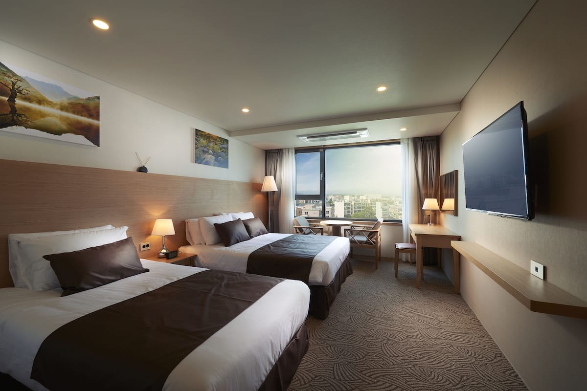 A hotel room with two beds, a desk, a chair, a TV, and a large window offering a city view.