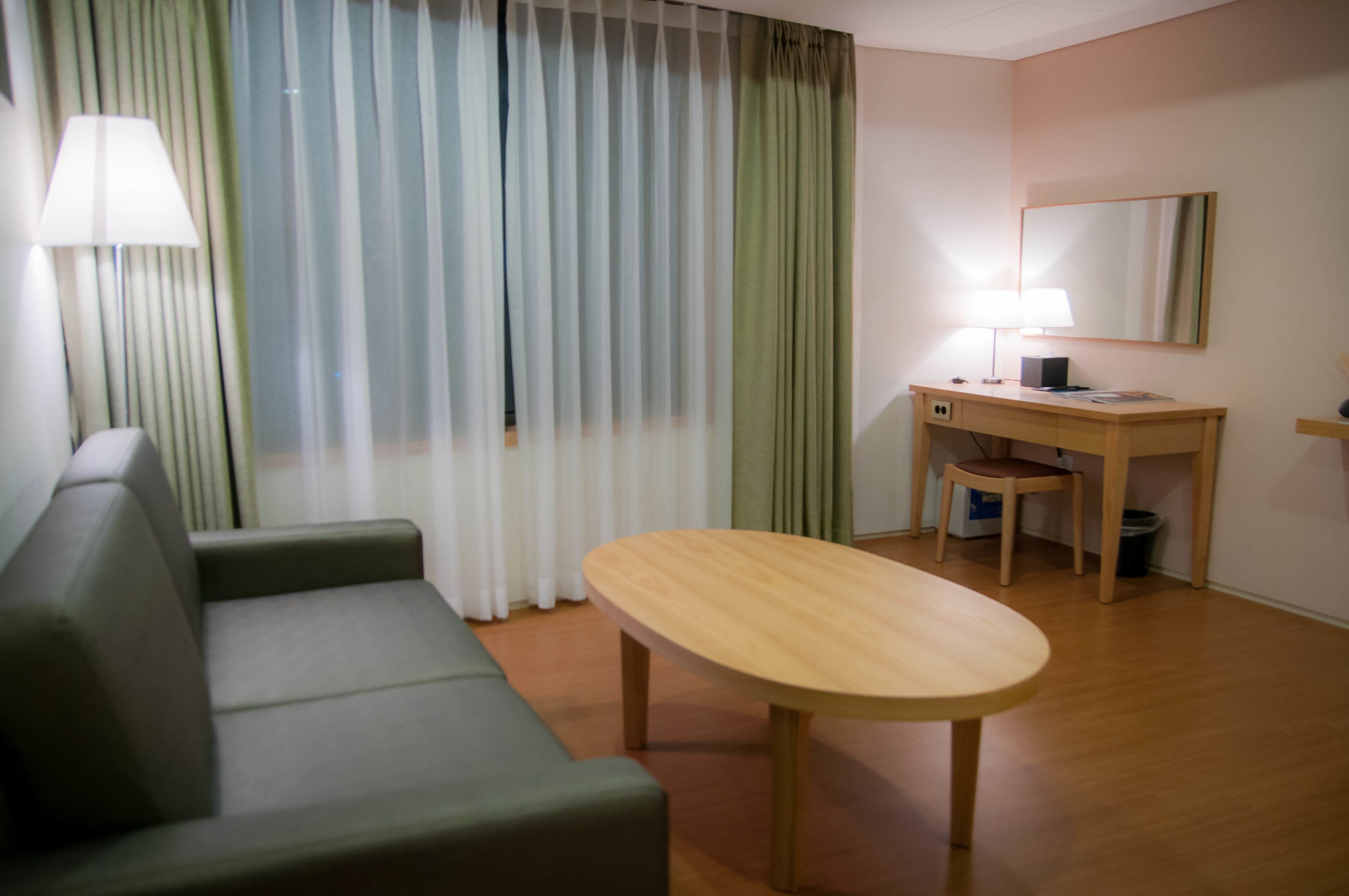 A hotel room with a green sofa, a wooden table, a desk with a chair, a lamp, and a large mirror on the wall.